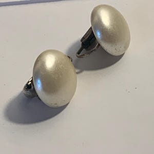 Puede incluir: Un par de pendientes de clip con grandes perlas ovaladas de color blanco roto. Los pendientes tienen un mecanismo de clip de metal plateado en la parte posterior. Las perlas tienen un brillo sutil, que refleja la luz.