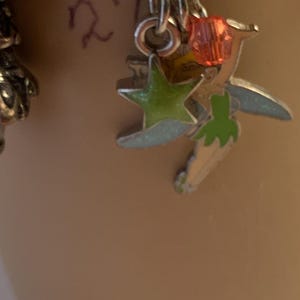 Puede incluir: Pendientes con colgantes plateados con una estrella verde, un hada y una cuenta roja. El hada tiene detalles verdes y blancos con alas azul claro. Los pendientes cuelgan de un aro plateado.