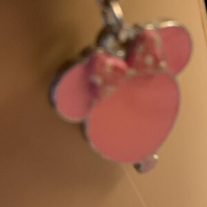 Puede incluir: Un charm rosa con un dise&ntilde;o de Minnie Mouse. El charm presenta un lazo rosa con lunares blancos y orejas rosas. El charm est&aacute; unido a una cadena plateada.