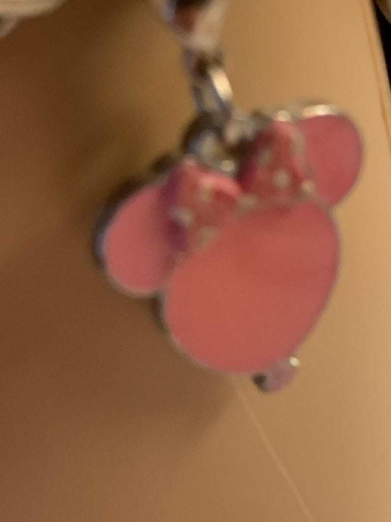 Puede incluir: Un charm rosa con un dise&ntilde;o de Minnie Mouse. El charm presenta un lazo rosa con lunares blancos y orejas rosas. El charm est&aacute; unido a una cadena plateada.