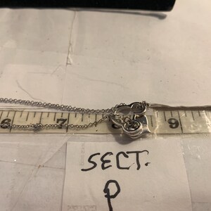 May include: A silver-toned necklace with a heart-shaped pendant. The pendant has a decorative design and small gemstones. The necklace is measured with a ruler, showing the chain length. A note reads "SECT. P".