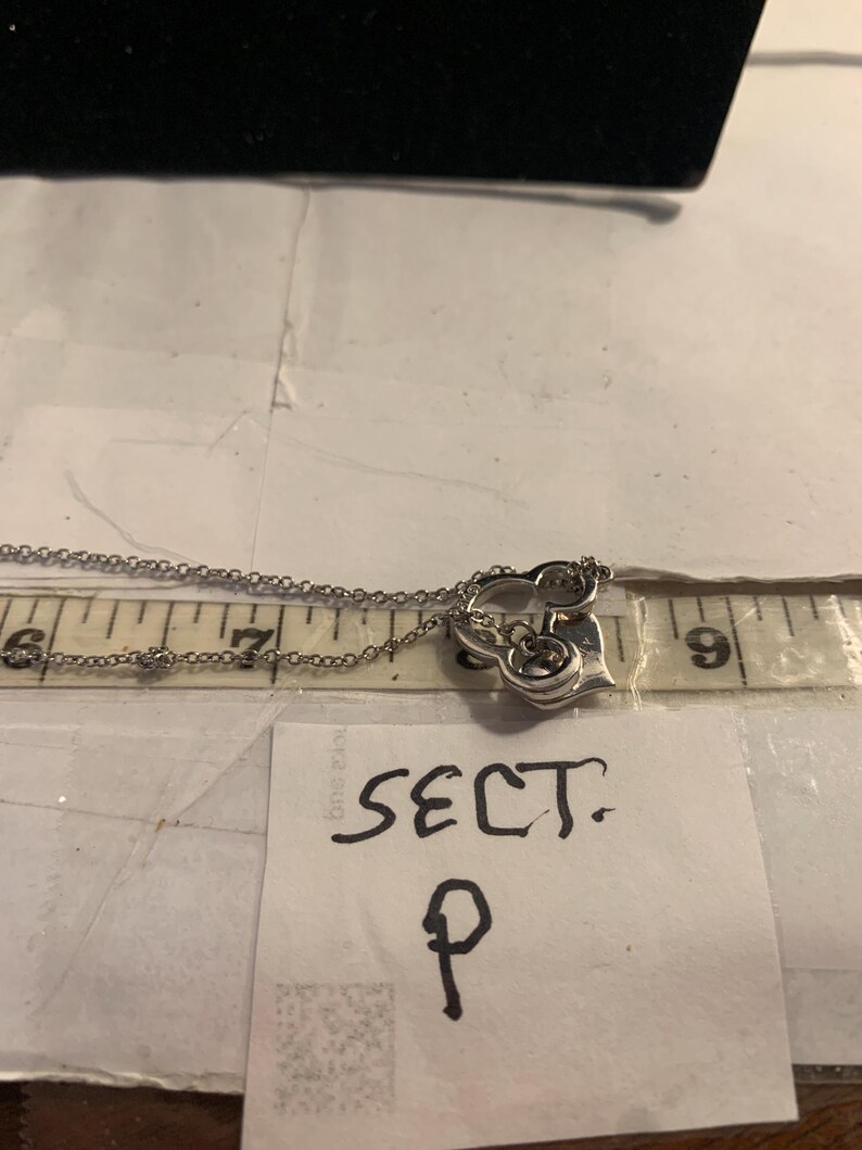 May include: A silver-toned necklace with a heart-shaped pendant. The pendant has a decorative design and small gemstones. The necklace is measured with a ruler, showing the chain length. A note reads "SECT. P".