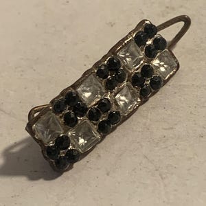 May include: A rectangular barrette with a metal frame, adorned with alternating clear square and black round gemstones. The barrette has a curved metal clasp on one end. The gemstones are set closely together, creating a sparkling effect.