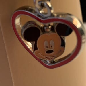 Puede incluir: Colgante plateado en forma de coraz&oacute;n con borde rojo. Dentro del coraz&oacute;n, una cara sonriente de Mickey Mouse con orejas negras y cara color beige. El colgante est&aacute; sujeto a una cadena plateada.