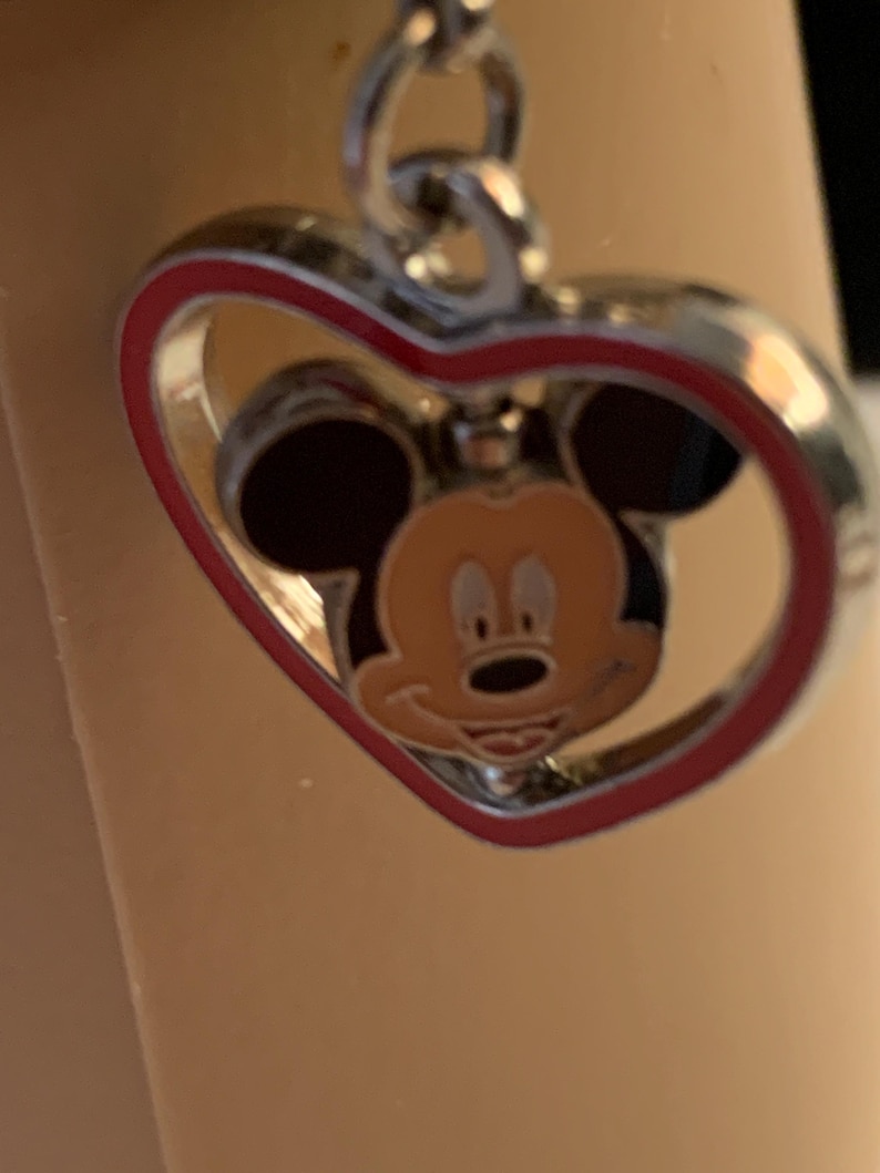 Puede incluir: Colgante plateado en forma de coraz&oacute;n con borde rojo. Dentro del coraz&oacute;n, una cara sonriente de Mickey Mouse con orejas negras y cara color beige. El colgante est&aacute; sujeto a una cadena plateada.