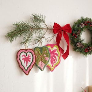 May include: Three heart-shaped tufted textile ornaments featuring leaf patterns in pink, green, and white, hanging from a pine branch decorated with a large red velvet bow and a small evergreen wreath with red berries and pinecones.