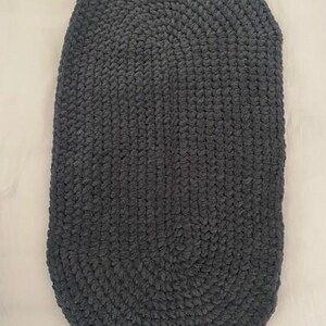 May include: A dark gray, oval-shaped, hand-crocheted rug. The rug is made of a thick, plush yarn, and the stitches are clearly visible. The rug's dimensions are approximately 30 inches long and 20 inches wide. The rug is suitable for a bathroom or bedroom.
