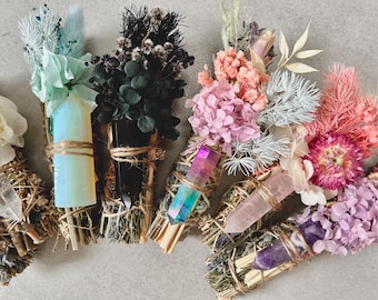 May include: A selection of sage bundles decorated with dried flowers and crystals. The bundles are in various colours, including pink, purple, blue, and black, and are tied with twine. The crystals are in various shapes and colours.
