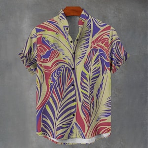 Puede incluir: Camisa de manga corta con estampado botánico. La camisa beige presenta grandes hojas estilizadas en morado, rojo y amarillo. Está abotonada y colgada de una percha de madera.