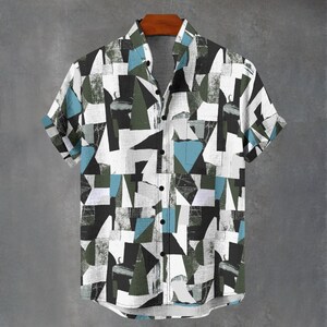 Puede incluir: Camisa de manga corta con un diseño geométrico en blanco, negro, gris, verde azulado y verde oliva. La camisa está abotonada y colgada de una percha de madera sobre un fondo gris.