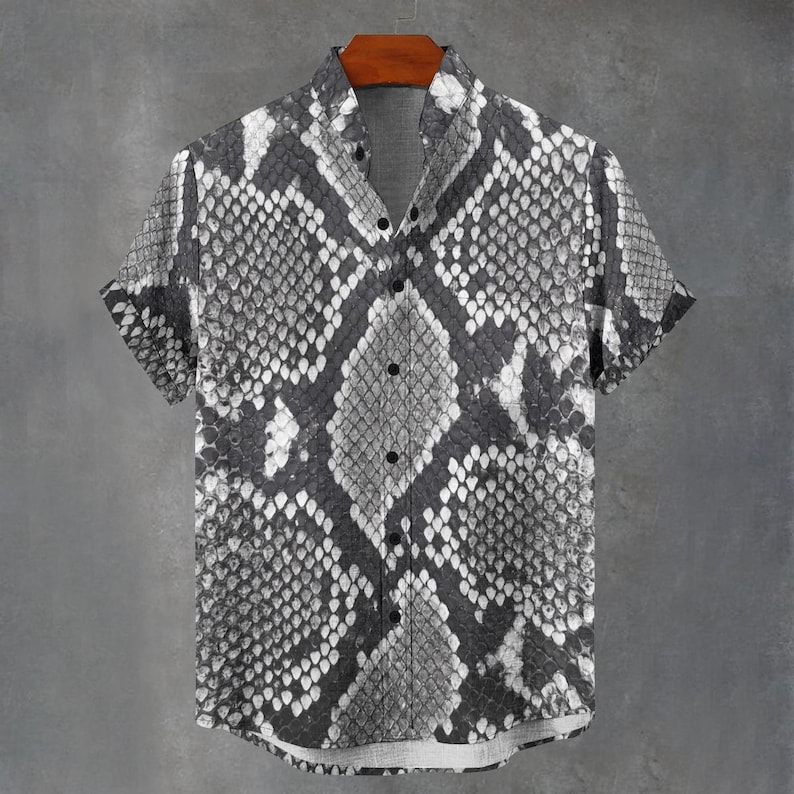 May include: A short-sleeved button-down shirt featuring a monochromatic snakeskin pattern in shades of black, white, and grey. The garment has a standard collar, a front button placket with dark buttons, and a straight hemline with cuffed sleeves.