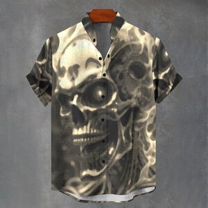 May include: Short-sleeved button-down shirt with a sepia-toned skull graphic. The shirt features a mandarin collar and black buttons. The skull design is prominently displayed across the front and back of the shirt, creating a bold, edgy look.