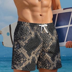 Puede incluir: Pantalones cortos de baño con estampado de serpiente en negro y beige con cordón blanco. Los pantalones cortos tienen un corte clásico por encima de la rodilla. El fondo muestra a una persona sosteniendo una tabla de surf.