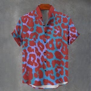 Puede incluir: Camisa de manga corta con un estampado de leopardo llamativo. La camisa tiene un cuello rojo y un bolsillo en el pecho. El patrón presenta formas rojas, azules y moradas sobre un fondo azul. Cuelga de una percha de madera.