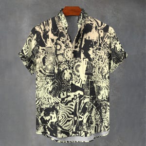 Puede incluir: Camisa de manga corta con un diseño abstracto en negro y crema. La camisa tiene cuello y un corte holgado. El patrón incluye texto y varias formas. La camisa está colgada en una percha de madera.