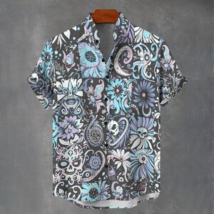 May include: Short-sleeved button-down shirt with a dark grey background and a repeating pattern of skulls, flowers, and paisley designs in shades of blue, purple, and white. The shirt is hanging on a wooden hanger.