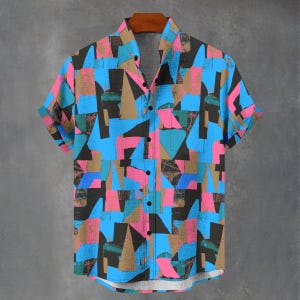 May include: Short-sleeved shirt with a bold geometric design in shades of blue, pink, brown, green, and black. The shirt is buttoned up and hangs on a wooden hanger. The background is a plain grey wall.