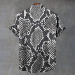 May include: A short-sleeved button-down shirt featuring a monochromatic snakeskin pattern in shades of black, grey, and white. The garment has a standard collar, a straight hemline, and a repeating scale texture across the entire surface.