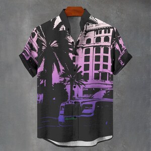 May include: Short-sleeved button-down shirt with a black, purple, and white graphic print. The design features palm trees, a building, and a car. The shirt has a classic collar and is hanging on a wooden hanger.