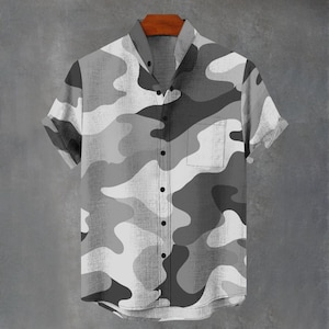 May include: Short-sleeved button-down shirt with a gray and white camouflage pattern. The shirt has a single chest pocket and a mandarin collar. The shirt is hanging on a wooden hanger against a gray background.
