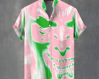 May include: Pink short-sleeved shirt with a green and white graphic design. The shirt has a button-down front and a collared neckline. The graphic appears to be a stylized face. The shirt is hanging on a wooden hanger.