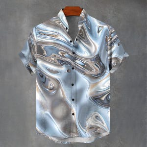 Pode incluir: Camisa de manga curta com um padrão abstrato em tons de azul, cinza e castanho. A camisa tem uma gola clássica, um bolso no peito e botões pretos. O tecido parece ter um efeito líquido ou marmoreado.