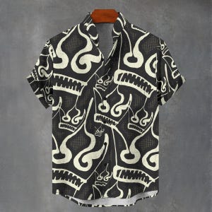 Puede incluir: Camisa negra y blanca de manga corta con un patrón único. La camisa presenta un diseño repetido de coronas estilizadas y formas de llama. La camisa está colgada en una percha de madera sobre un fondo gris.