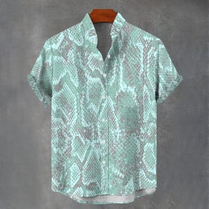 Può includere: Una camicia a maniche corte con un motivo a pelle di serpente nei toni del verde acqua e grigio. La camicia ha un colletto alla coreana e una tasca sul petto. Le maniche sono risvoltate. La camicia è esposta su una gruccia di legno.