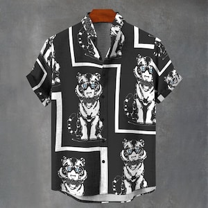 May include: Black button-down shirt with a repeating pattern of white tiger illustrations. The tigers have blue eyes and are framed by white geometric shapes. The short-sleeved shirt hangs on a wooden hanger against a gray background.