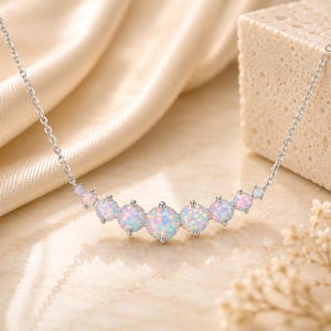 May include: A silver-toned necklace featuring a curved row of seven round, iridescent opal-like stones that graduate in size, with the largest stone positioned in the center. The stones display flashes of pink, blue, and white light.
