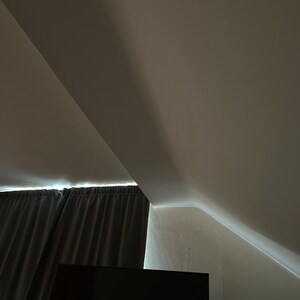 May include: A dark grey curtain hangs in front of a window, with a black television screen below. The room has a sloping ceiling with white walls.