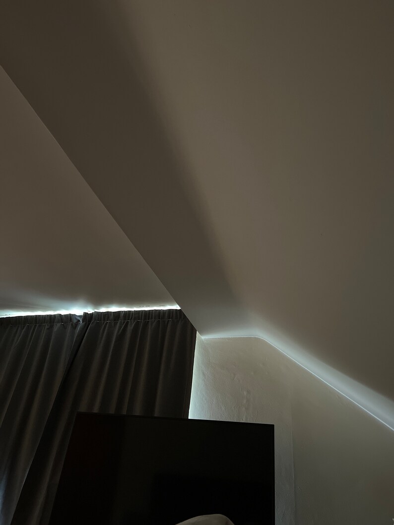 May include: A dark grey curtain hangs in front of a window, with a black television screen below. The room has a sloping ceiling with white walls.