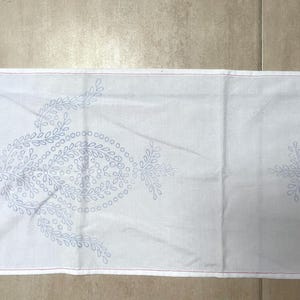 May include: White fabric table runner with a delicate blue embroidered design of repeating floral patterns.