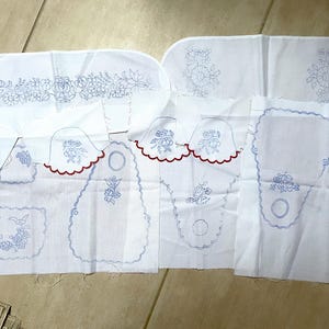 Puede incluir: Una colección de piezas de tela blanca con diseños de bordado azul. Las piezas son de varias formas y tamaños, incluyendo rectángulos, óvalos y círculos. Algunas piezas tienen ribetes rojos.