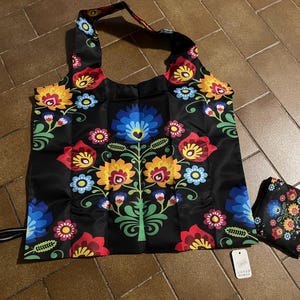 May include: Black tote bag with a colorful floral design. The bag features a repeating pattern of red, yellow, blue, and pink flowers with green leaves. A smaller, matching bag is folded next to it. The bag has a black handle.