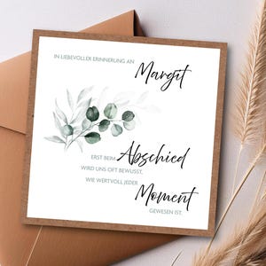 Könnte beinhalten: Eine Beileidskarte mit braunem Rand und Umschlag. Die Karte zeigt den Namen "Margit" in eleganter Schrift sowie die Worte "Abschied" und "Moment". Ein Eukalyptuszweig im Aquarellstil verleiht dem Design eine natürliche Note.