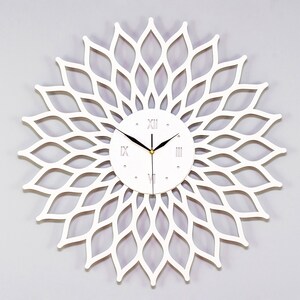 Könnte beinhalten: Weiße Wanduhr mit einem dekorativen, filigranen Blumendesign. Das Zifferblatt der Uhr hat römische Ziffern und schwarze Zeiger.