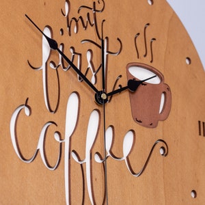 Op de afbeelding: Een houten klok met een koffiekopontwerp en de tekst "but first, coffee". De klok heeft zwarte wijzers en een bruine achtergrond.
