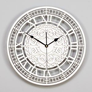 May include: White wall clock with Roman numeral markings and a decorative, intricate, white, cutout design in the centre. The clock hands are black.