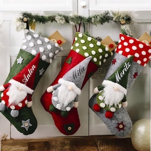 Op de afbeelding: Vier kerstsokken met kabouterdecoraties. De sokken zijn rood, groen en grijs met witte stippen. Elke sok heeft een naam: Anton, Mila en Moritz. Een feestelijke vakantiedecoratie.