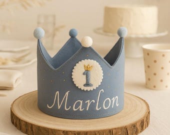 Op de afbeelding: Een stoffige blauwe vilten kroon met een witte geschulpte ronde patch met een blauw cijfer één en een klein gouden kroonicoontje. De naam Marlon is in wit schrift geborduurd. De kroon is afgewerkt met kleine blauwe en witte pompons.