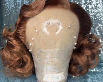 May include: A copper-colored wig with voluminous curls styled on a mannequin head. The wig features a lace front cap secured with white pearl-headed pins. A small white label with the handwritten name Amy is attached to the base of the head.