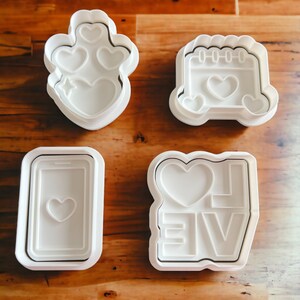 May include: Four white plastic cookie cutters shaped like a heart with a star, a calendar with a heart, a phone with a heart, and the word "LOVE" with a heart.