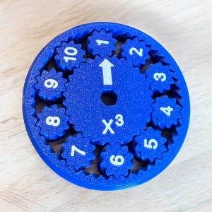 May include: A blue gear-shaped educational toy with white numbers 1-10 and an arrow. The center has the text "X³". The toy is designed with interlocking gears and is likely used for teaching math concepts.