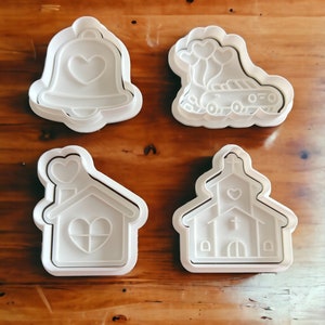 May include: Four white plastic cookie cutters, shaped like a bell with a heart, a house with a heart, a church with a cross and a heart, and a car with balloons in the shape of hearts.