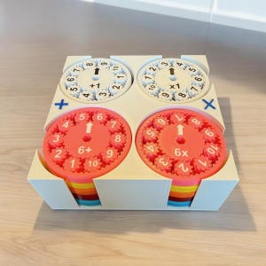 May include: A white plastic box with four colorful circular number wheels. The wheels have numbers 1-10 and symbols for addition, subtraction, multiplication, and division. The wheels are designed for learning basic math.