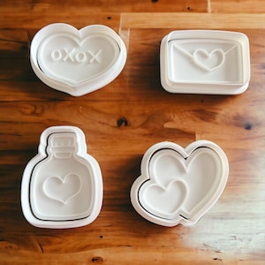 May include: Four white plastic cookie cutters on a wooden surface. The cutters are shaped like a heart with the word "oxox" inside, a heart inside an envelope, a bottle with a heart inside, and two hearts nested inside each other.