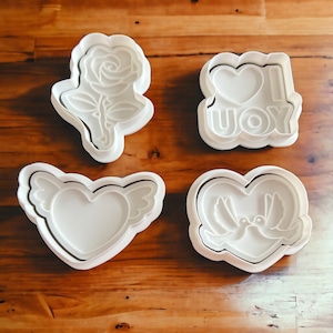 May include: Four white plastic cookie cutters on a wooden surface. The cutters are shaped like a rose, a heart with wings, a heart with doves, and a heart with the words "I love you".