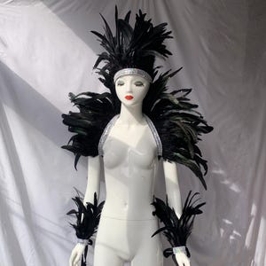 May include: A mannequin wearing a black feather costume. The costume features a feathered headdress, shoulder piece, arm cuffs, and a headband, all embellished with silver rhinestones. The feathers are a dark black.
