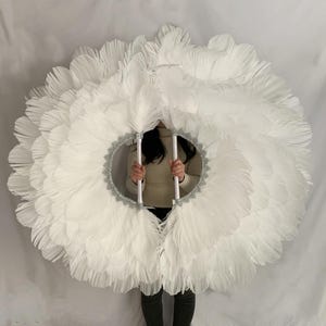 May include: A large, circular white feather fan with a silver glitter trim and two silver handles. The fan is held in front of a person, creating a dramatic effect. The feathers are layered, giving the fan a full, fluffy appearance.